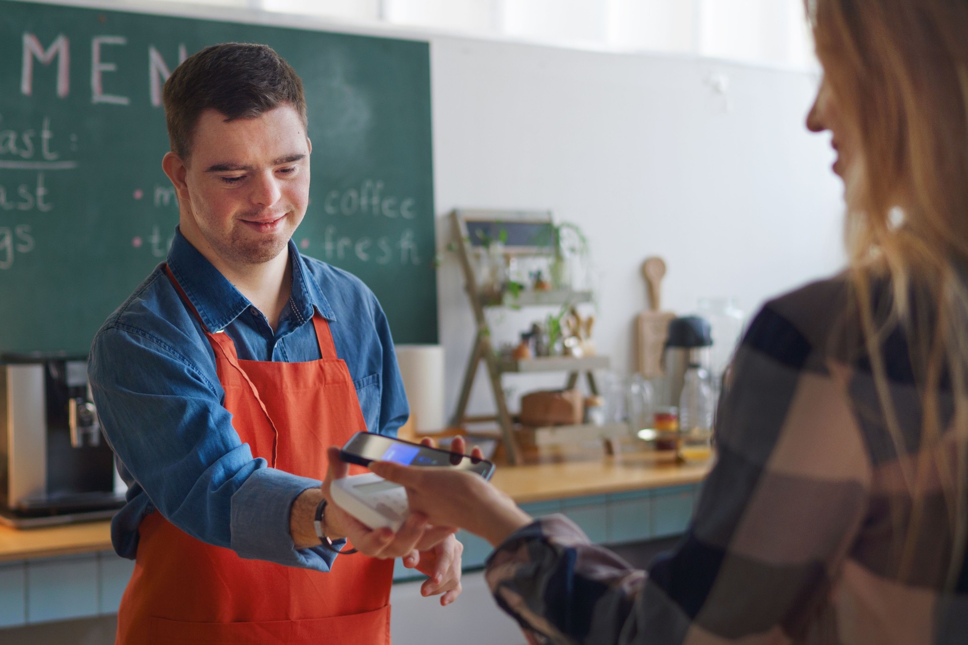  Cheerful young man with Down syndrome working as a waiter, taking contactless smartphone payment from a customer—representing inclusive employment, independence, and workforce participation for adults with developmental disabilities